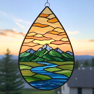 Pode incluir: Um apanhador de sol em forma de lágrima de vidro colorido que representa uma cordilheira com um rio serpenteando por um vale. O céu é um pôr do sol laranja e rosa vibrante com nuvens.