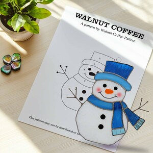 May include: A stained glass snowman ornament with a blue hat, scarf, and twig arms. The snowman has a carrot nose and rosy cheeks. The ornament is displayed on a white paper with the text "WALNUT COFFEE".
