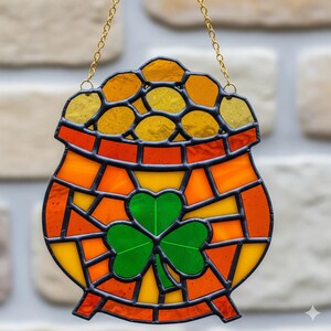 May include: Stained glass pot of gold with a green shamrock. The pot is orange and yellow with a black outline, and is hanging from a gold chain. The background is a brick wall.