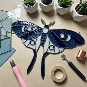 Puede incluir: Una vidriera con forma de polilla en tonos azules y blancos, con detalles de luna creciente. La imagen incluye herramientas para trabajar el vidrio y pequeñas suculentas en macetas. La polilla tiene un contorno azul oscuro y está hecha de vidrio azul y blanco.