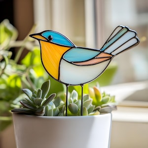 May include: A stained glass bird with blue, yellow, and white accents, perched on a metal stand. The bird is in front of a potted succulent plant.