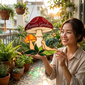May include: Stained glass garden sign featuring a red mushroom with white spots and the text "Anna's Garden." The sign also includes a smaller orange mushroom, a snail, and green leaves. The sign is hanging from a chain.