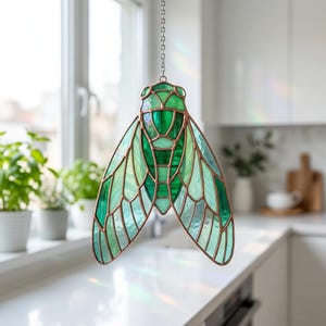 May include: A stained glass insect decoration with shades of green and copper-colored metal framing. The decorative piece is hanging from a chain and is in the shape of a cicada. The wings are a mosaic of green glass.
