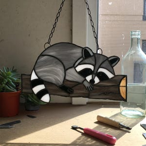 May include: A stained glass window hanging with a raccoon sleeping on a tree branch. The raccoon is grey with black and white stripes on its tail. The tree branch is brown with a light brown centre.