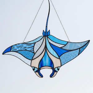 May include: Stained glass manta ray in shades of blue, white, and cream, with a textured and patterned design. The artwork is suspended by chains, showcasing the aquatic theme. The ray's wingspan is approximately 20 inches.