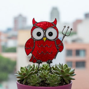 May include: A red stained glass owl with devil horns and a trident, perched above a succulent plant in a purple pot. The owl has large white eyes and a yellow beak. The background is blurred, showing a cityscape.