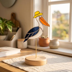 May include: A stained glass pelican sculpture with a yellow hat, orange beak, and blue and white body. The bird is mounted on a gold-colored metal rod and a round wooden base. The sculpture is displayed on a table near a window.
