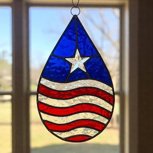 Könnte beinhalten: Ein tropfenförmiger Anhänger aus Buntglas mit einem rot-weiß-blauen amerikanischen Flaggenmuster. Der Anhänger zeigt einen weißen Stern auf blauem Hintergrund und rote und weiße Streifen.