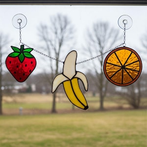 May include: Stained glass fruit garland featuring a red strawberry, a yellow banana, and an orange slice. Each piece is outlined in black and hangs from a chain with suction cups for window display.