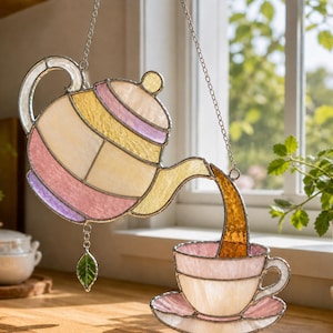 Puede incluir: Vidriera de una tetera vertiendo té en una taza. El diseño presenta tonos rosas, amarillos y blancos, con un dije de hoja verde. El té se representa de color ámbar, creando una estética cálida.