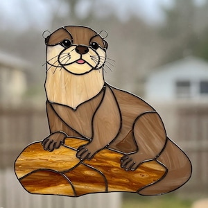 Puede incluir: Una ventana de vitral colgante que presenta una nutria marr&oacute;n y tostada sentada en una roca. La nutria tiene ojos negros y una nariz rosa.