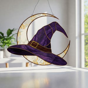 May include: A stained glass witch hat in shades of purple and brown, with a gold buckle, hangs in front of a crescent moon. The moon is white with gold accents and is suspended by a chain, creating a whimsical Halloween decoration.