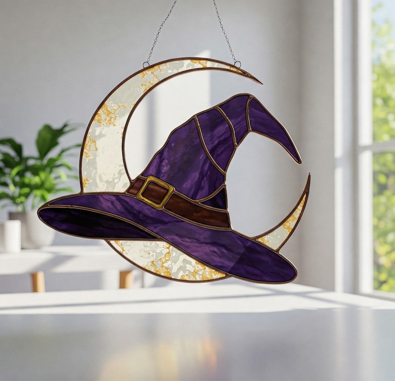May include: A stained glass witch hat in shades of purple and brown, with a gold buckle, hangs in front of a crescent moon. The moon is white with gold accents and is suspended by a chain, creating a whimsical Halloween decoration.
