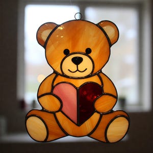 May include: Stained glass teddy bear holding a heart. The bear is a warm amber colour with black accents and a smiling face. The heart is divided into pink and red sections, creating a charming decorative piece.