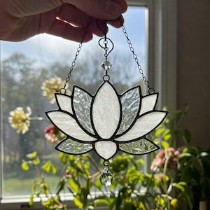 May include: A stained glass lotus flower suncatcher with a black metal frame and chain. The flower has white, iridescent petals and a clear crystal bead at the bottom. The suncatcher is hanging in front of a window.