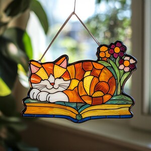 May include: A stained glass window hanging features a sleeping orange and white cat curled up on a yellow book with green leaves and pink, red, and white flowers.