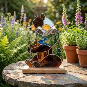 May include: A stained glass bear figure standing on a light wood base. The bear features a landscape scene within its body, including a yellow sun, white mountains, a blue river, and green pine trees. The bear holds a small green pine tree.