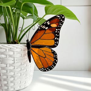 May include: A stained glass butterfly with orange, black, and yellow wings. The butterfly is perched on a white wicker planter with a green plant.