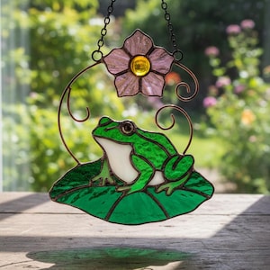 May include: Stained glass frog suncatcher with a green frog on a lily pad, a pink flower, and a bronze-colored metal frame. The suncatcher hangs from a chain and is displayed in front of a blurred outdoor background.