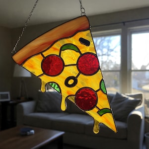 Puede incluir: Una rebanada de pizza de vidrio coloreado que cuelga de una cadena. La pizza es amarilla con pepperoni rojo, pimientos verdes y una aceituna negra. La rebanada gotea con queso derretido.