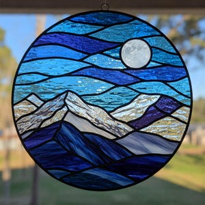May include: A round stained glass panel depicting a mountain landscape under a full moon. The design features shades of blue, white, and purple, with the moon rendered in white. The panel is designed to be hung.
