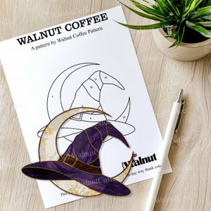 May include: A printed pattern for a stained glass project featuring a witch's hat and crescent moon design. The pattern is labeled "WALNUT COFFEE" and includes the text "A pattern by Walnut Coffee Pattern". A white pen rests on the wooden surface.