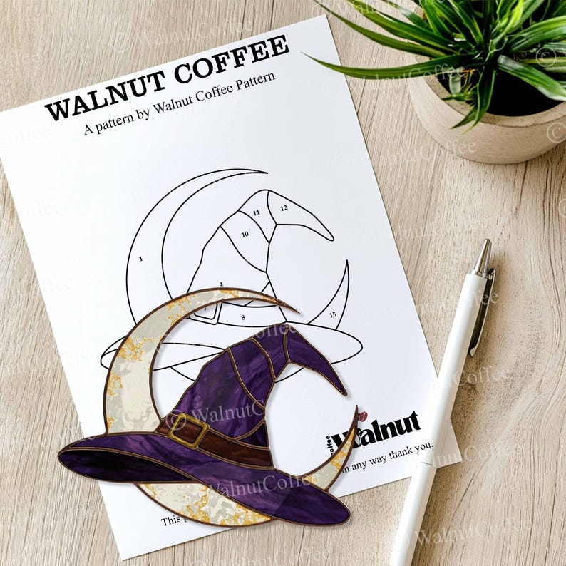 May include: A printed pattern for a stained glass project featuring a witch's hat and crescent moon design. The pattern is labeled "WALNUT COFFEE" and includes the text "A pattern by Walnut Coffee Pattern". A white pen rests on the wooden surface.