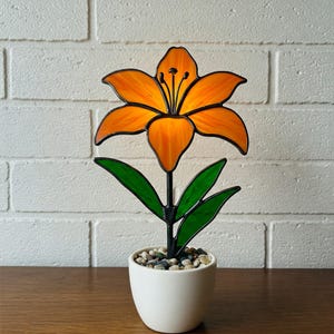 Puede incluir: Una flor de lirio de vidrio emplomado en naranja y verde, con un tallo y hojas de metal negro. La flor está sentada en una maceta de cerámica blanca con pequeñas piedras.