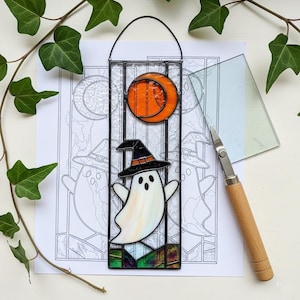 Op de afbeelding: Een gebrandschilderde Halloween-decoratie met een spook met een heksenhoed, een oranje maan en een zwart frame. De decoratie hangt aan een zwarte draad. Een knutselmes en een tekening staan op de achtergrond.