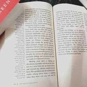 Puede incluir: Un libro de tapa dura abierto titulado The Art of Seduction de Robert Greene, con texto impreso en negro sobre p&aacute;ginas blancas. El libro est&aacute; colocado junto a un libro cerrado con una cubierta roja y azul, mostrando el nombre del autor en blanco.