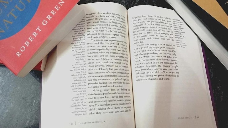 Puede incluir: Un libro de tapa dura abierto titulado The Art of Seduction de Robert Greene, con texto impreso en negro sobre p&aacute;ginas blancas. El libro est&aacute; colocado junto a un libro cerrado con una cubierta roja y azul, mostrando el nombre del autor en blanco.