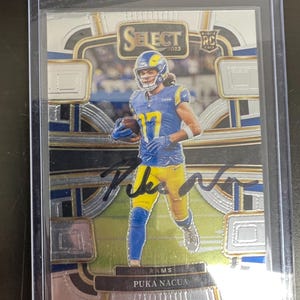 Puede incluir: Una tarjeta de fútbol RC Select 2023 con Puka Nacua con el uniforme de Los Angeles Rams. La tarjeta está dentro de un soporte protector e incluye un autógrafo. La tarjeta tiene un diseño plateado, azul y dorado.