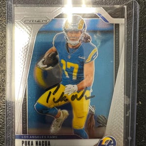 Può includere: Una carta collezionabile di football di Puka Nacua dei Los Angeles Rams. La carta presenta un giocatore in uniforme blu e giallo, che tiene un pallone da football. La carta è racchiusa in una custodia protettiva. La carta è firmata.
