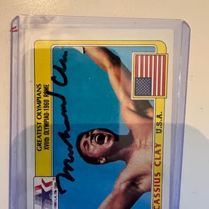 May include: A trading card featuring Cassius Clay, with his signature in blue ink. The card has a yellow border and includes text that reads "Greatest Olympians XVIIth Olympiad-1960 Rome" and "U.S.A."