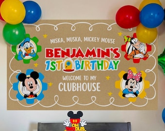 May include: A birthday party setup featuring a large banner with Mickey Mouse characters and the text 'Benjamin's 1st Birthday Welcome to My Clubhouse', a white cake with a Mickey topper, popcorn containers, and colorful balloons in red, yellow, blue, and green.