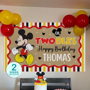 Puede incluir: Un telón de fondo de cumpleaños con Mickey Mouse, el texto "OH, TWODLES Happy Birthday THOMAS" y un esquema de colores rojo, amarillo, negro y blanco. Un pastel con un adorno a juego, recipientes de palomitas y globos decoran la escena.