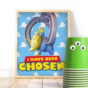 May include: A framed poster featuring a green three-eyed alien character being lifted by a mechanical claw against a blue sky with white clouds, with the text I HAVE BEEN CHOSEN in red and yellow, next to a stack of green cups with three eyes.