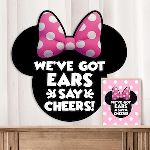 Könnte beinhalten: Ein schwarzes mausförmiges Schild mit einer rosa-weiß gepunkteten Schleife, das den weißen Text WE'VE GOT EARS SAY CHEERS! und zwei weiße Handschuhsymbole zeigt. Daneben steht ein gerahmter Druck mit demselben Motiv auf einem rosa gepunkteten Hintergrund.