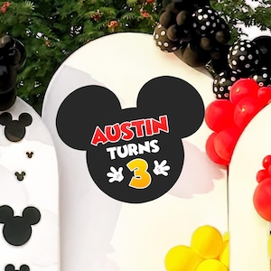 Peut inclure: Un panneau noir en forme de tête de Mickey Mouse avec le texte "Austin Turns 3" en rouge et jaune. Le panneau fait partie d'une décoration de fête d'anniversaire, entouré de ballons rouges, jaunes et noirs. L'arrière-plan est une arche blanche.
