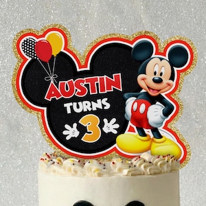 May include: A white frosted cake decorated with a black Mickey Mouse silhouette and a colorful border of red, yellow, and black circles. A glittery gold-edged cake topper features Mickey Mouse, three balloons, and the text AUSTIN TURNS 3.