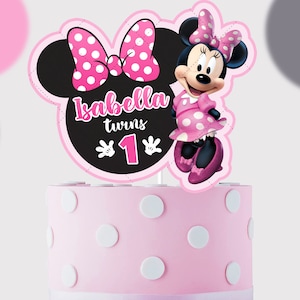 May include: A pink cake topper featuring Minnie Mouse, a large black Mickey Mouse ear silhouette with a pink polka dot bow, and the text Isabella turns 1. The topper sits on a light pink cake decorated with white polka dots and a white ribbon base.