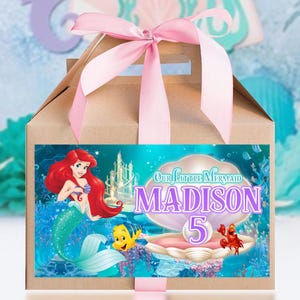 May include: A brown cardboard gable box with a pink satin ribbon tied at the top. The front features a label with a mermaid, a yellow fish, a red crab, and a large seashell containing the text Our Little Mermaid, Madison, and the number 5.