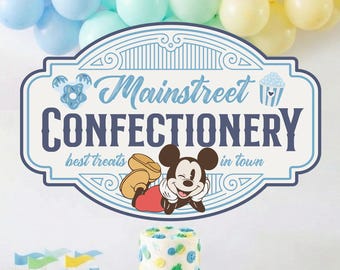 May include: A party table featuring a cake, bowls of treats, and a sign reading Mainstreet CONFECTIONERY best treats in town with a Mickey Mouse illustration. Above the sign are blue, mint, and yellow balloons. A Disneyland sign is also present.