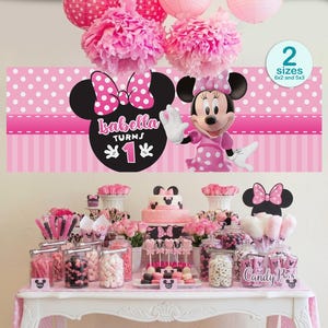 Op de afbeelding: Een roze Minnie Mouse themafeestopstelling. De tafel is versierd met een roze tafelkleed, lekkernijen en een taart. Een banner met Minnie Mouse en de tekst "Isabella Turns 1" is boven de tafel te zien. Roze pompons en papieren lantaarns hangen aan het plafond.