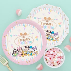 以下が含まれることがあります： ディズニーのキャラクターと「Elizabeth's Baby Shower」の文字が入った、ピンクと白のベビーシャワー用プレート。マシュマロの入ったボウルと金のフォークも写っています。プレートはパステルカラーです。