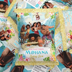 May include: A collection of rectangular party favor bags featuring a Moana theme with gold glitter edges. The bags display characters from the film, tropical flowers, and text that reads Moana turns two, Our Wayfinder, and Thank you for coming to my adventure.