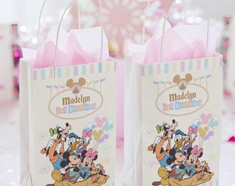May include: Two white party favor bags with clear handles, featuring Disney characters like Mickey and Minnie Mouse. Each bag has a pastel striped border, the name "Madelyn" and "1st Birthday" printed on the front, and is filled with pink tissue paper.