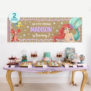 K&ouml;nnte beinhalten: Ein Banner mit dem Text "Our Little Mermaid Madison is turning 2" und Illustrationen zum Thema Meerjungfrau. Das Banner ist braun mit lila und wei&szlig;en Akzenten. Ein Tisch ist mit Desserts und Dekorationen gedeckt.