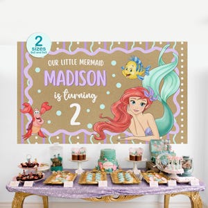 K&ouml;nnte beinhalten: Ein Geburtstagsbanner mit der kleinen Meerjungfrau Arielle, mit dem Text "Our Little Mermaid Madison is turning 2." Das Banner hat einen braunen Hintergrund mit lila und t&uuml;rkisfarbenen Akzenten. Ein Tisch ist mit Desserts gedeckt.