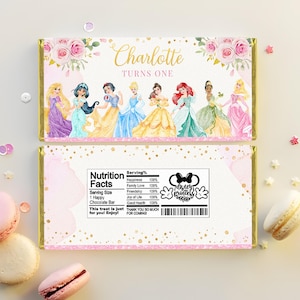 Könnte beinhalten: Rosa-goldene Schokoladentafel-Verpackung mit Blumenmuster und einem Bild von Disney-Prinzessinnen. Die Verpackung lautet "Charlotte wird eins" und enthält eine Nährwerttabelle mit einem Mickey-Maus-Symbol, auf dem "Disney Princess" steht.